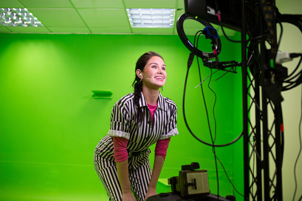 A woman in front of a green screen smiles into a camera and ring light.