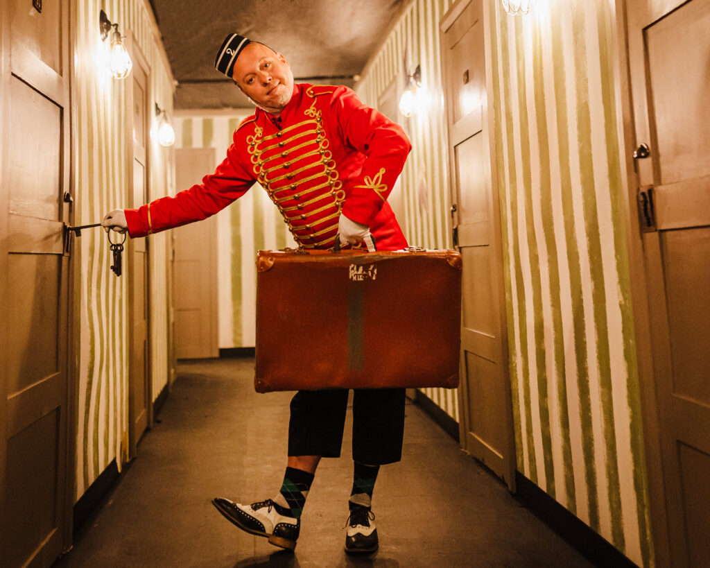 A man dressed in a bright red bell-boy outfit holds a suitcase and leans to the side opening a door with a key.